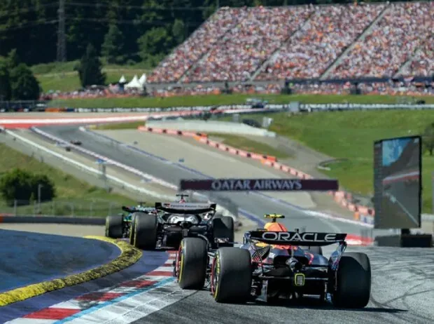 Several Formula 1 cars race closely uphill on the Red Bull Ring, with a gigantic, fully packed spectator grandstand visible in the background.