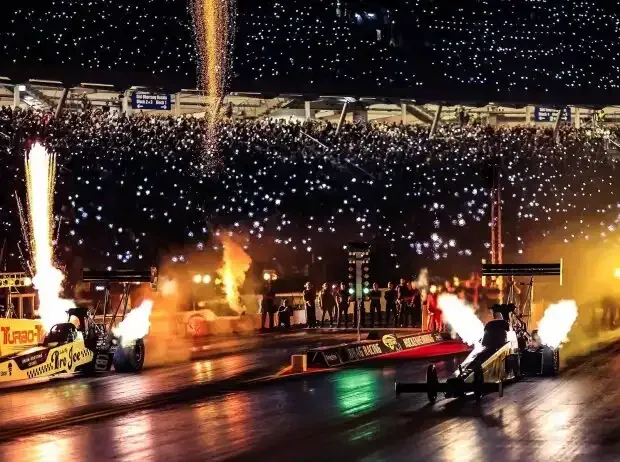 Title image for the news: Two dragsters stand at night on the starting line of a drag racing track and shoot huge flames from their exhaust pipes. In the background, the mobile phone lights of the fully occupied grandstand shine, while pyrotechnic sparks rain from the sky