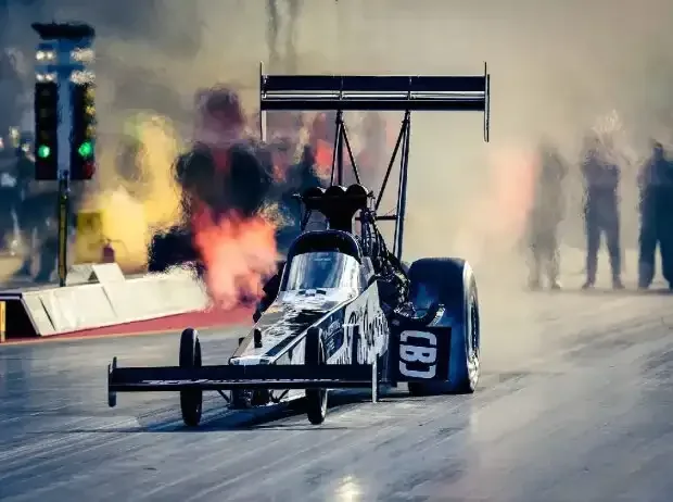 Title image for the news: A Top Fuel Dragster stands at the NitrOlympX drag racing event at the Hockenheimring surrounded by dense tire smoke and heat shimmer on the racetrack.